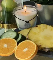Vela repelente estética junto a rodajas de naranja, pepino y piña sobre una mesa exterior.